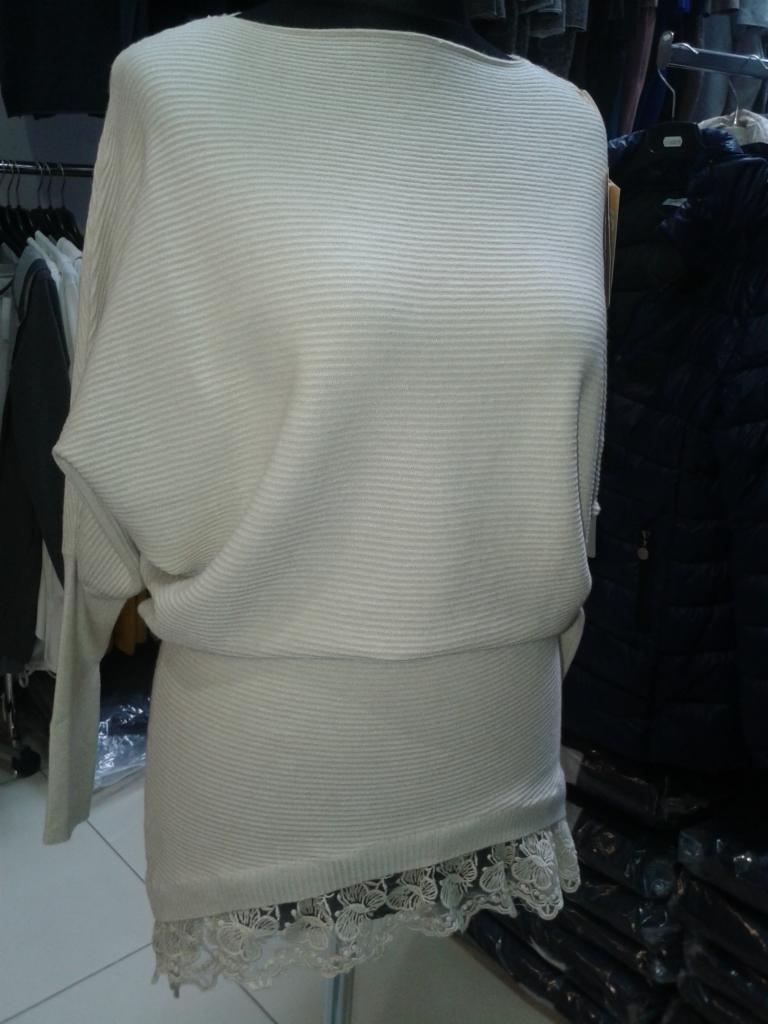 Kremowa tunika z dzianiny prążkowanej, z długim rękawem typu kimono i koronkowym wykończeniem dołu, prezentowana na manekinie na tle wieszaków z ubraniami.