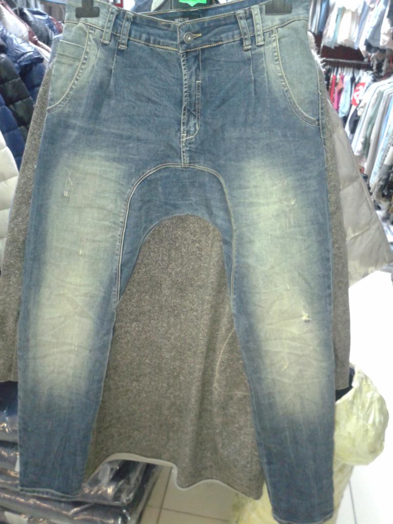 Niebieskie jeansy damskie o kroju baggy z niskim stanem, wiszące na wieszaku. Widoczne przetarcia i jasne przebarwienia na denimie. W tle inne ubrania.