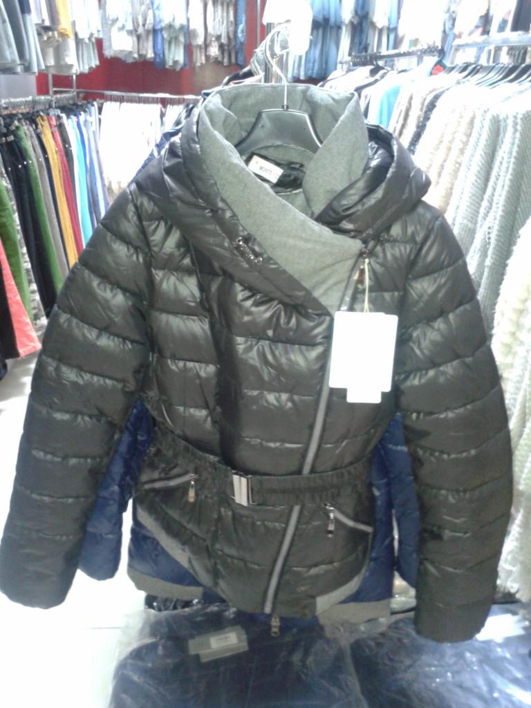 Czarna, pikowana kurtka damska z asymetrycznym zapięciem i szarym, wysokim kołnierzem, wisząca na wieszaku w sklepie odzieżowym. Widoczne inne ubrania w tle.