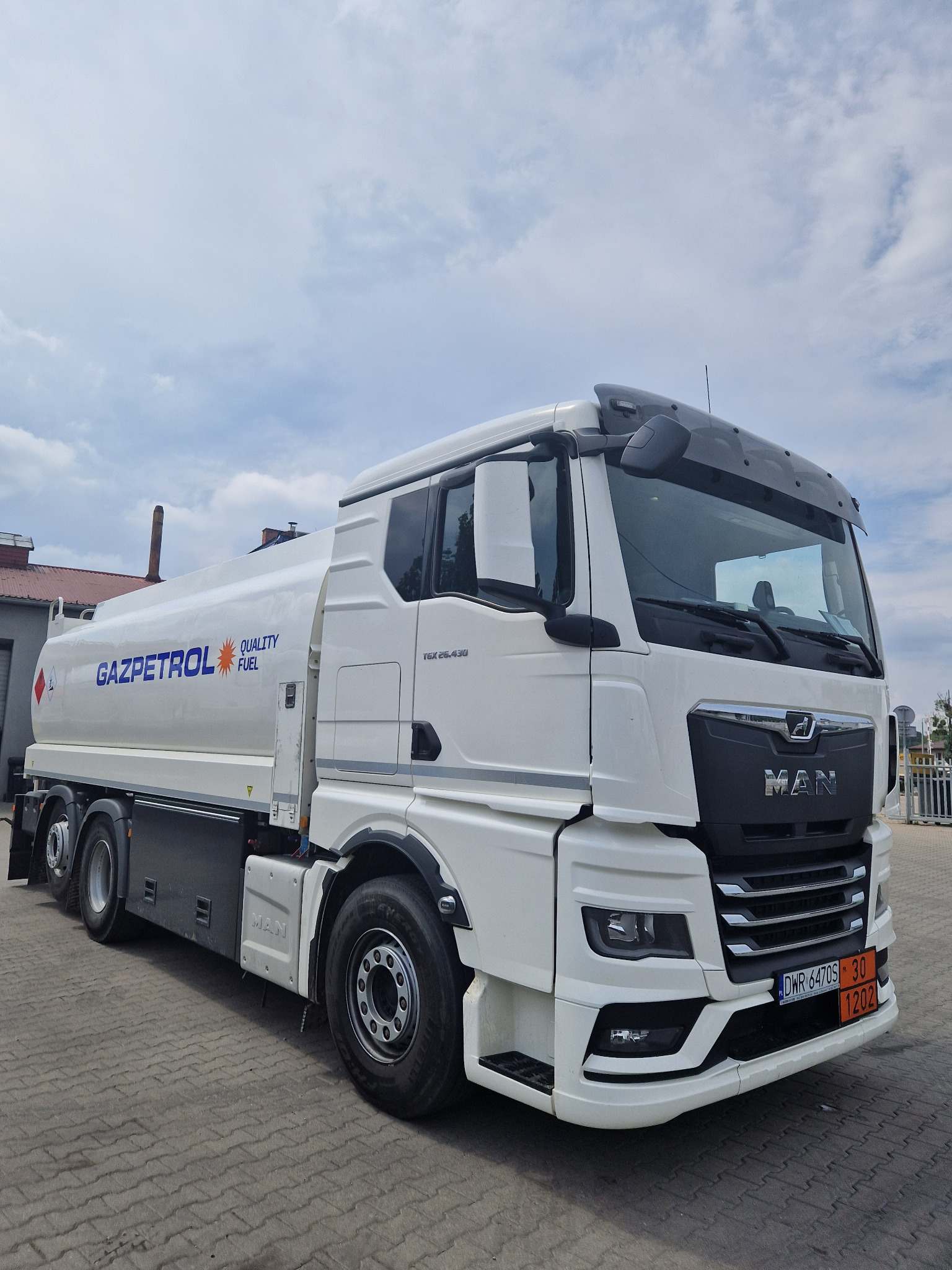Cysterna MAN Gazpetrol z oznaczeniami UN 1202 na parkingu, widok z przodu i boku, błękitne niebo w tle.