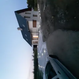 Nowoczesny dom jednorodzinny z podświetlonym podjazdem o zmierzchu, widok z dołu, częściowo widoczny samochód na pierwszym planie.