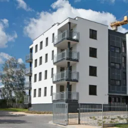 Nowoczesny, biały budynek wielorodzinny z balkonami o szklanych balustradach, widoczny zza metalowego ogrodzenia, na tle błękitnego nieba z chmurami i żurawiem budowlanym.