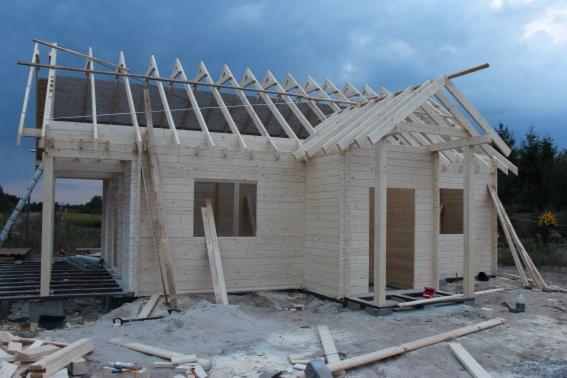 Konstrukcja drewnianego domu w trakcie budowy: widoczne ściany z bali, elementy więźby dachowej i fundamenty. Na pierwszym planie piasek i deski.