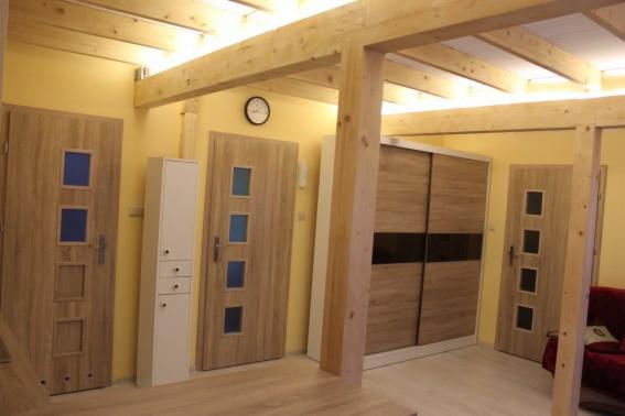 Wnętrze domu szkieletowego z drewnianą konstrukcją stropu, widoczne drzwi z szybkami, szafa przesuwna i fragment sofy. Żółte ściany, zegar na ścianie.