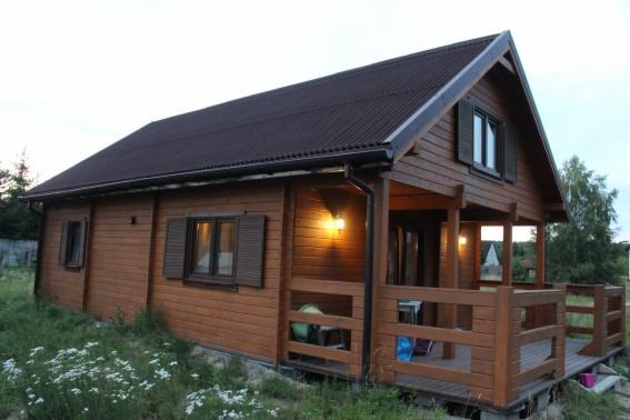 Drewniany dom z bali z brązowym dachem i oświetlonym gankiem, otoczony zielenią. Widoczne okiennice i elementy drewnianej konstrukcji.