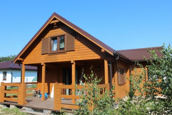 Gotowy dom z bali o brązowej elewacji i dachem, z widocznym tarasem, drewnianymi okiennicami i rynną. Błękitne niebo w tle.