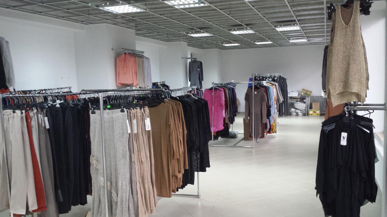 Wnętrze sklepu z odzieżą damską: rzędy ubrań na wieszakach, głównie spodnie i sukienki w stonowanych kolorach, oświetlone jarzeniówkami, widok w głąb sklepu.