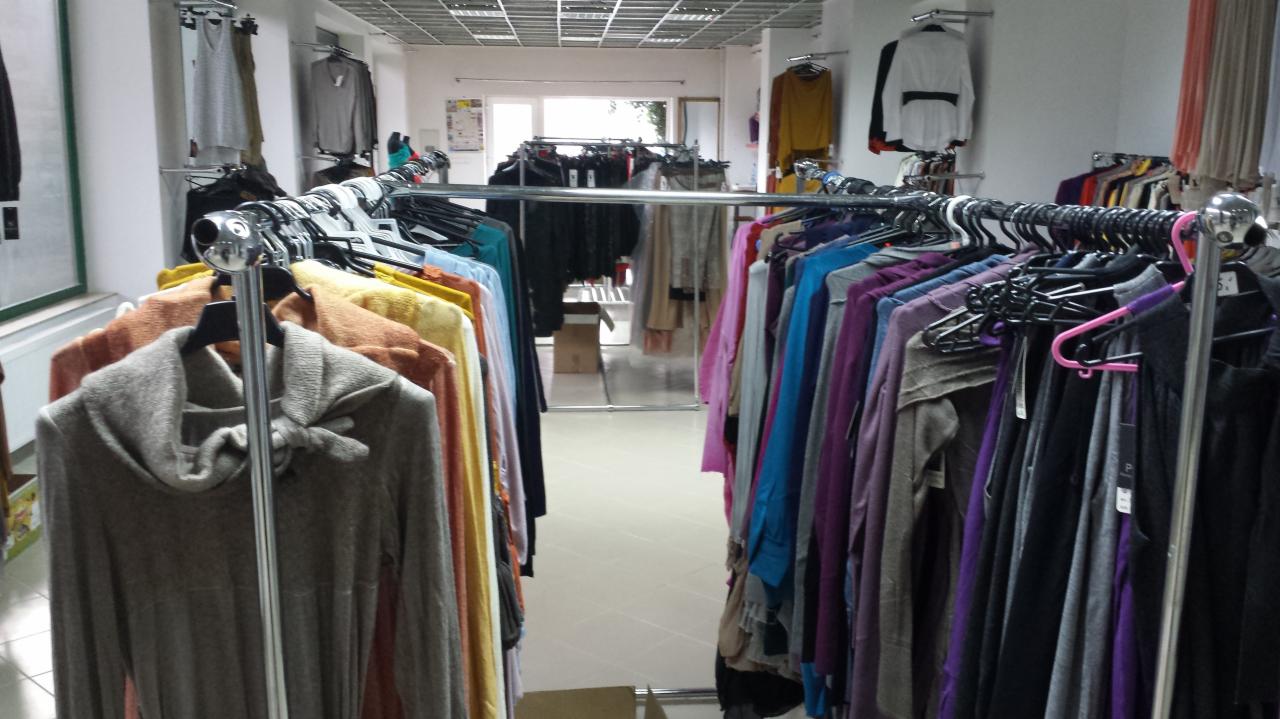 Wnętrze butiku odzieżowego z rzędami ubrań na wieszakach. Widoczne swetry, bluzki i sukienki w różnych kolorach. W tle okno i dalsze wieszaki z odzieżą.