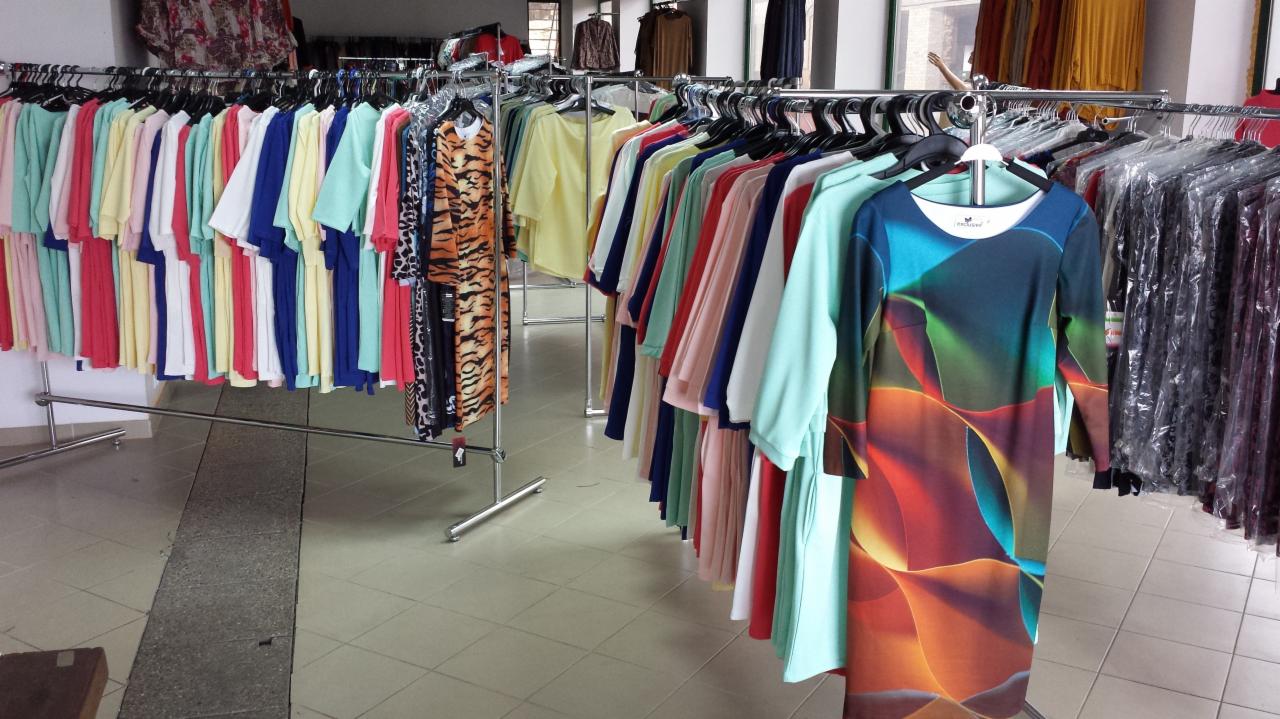 Wnętrze butiku z damską odzieżą. Na wieszakach wiszą kolorowe sukienki i bluzki, niektóre zapakowane w foliowe pokrowce. W tle widoczne inne ubrania.