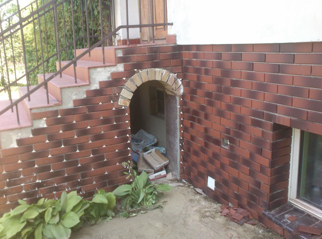 Wykończenie elewacji cegłą klinkierową w Kruszwicy, widoczne schody i łukowe wejście do piwnicy w trakcie prac. Detale mocowania płytek.