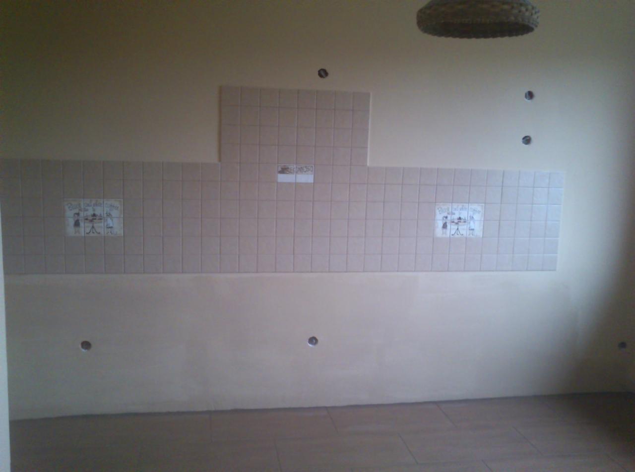 Fragment ściany z beżowymi płytkami w kuchni, z dekoracyjnymi motywami. Widoczne również otwory na gniazdka elektryczne. W tle widoczny fragment sufitu z lampą.