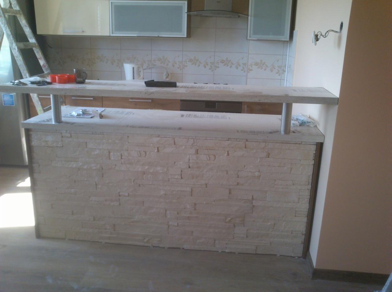 Wykończony barek kuchenny z dekoracyjną okładziną imitującą kamień, widok na fragment kuchni z białymi szafkami i drabina w tle.