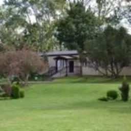 Parterowy budynek z ciemnym dachem i wejściem z rampą, otoczony zielonym trawnikiem i drzewami. Widok na ośrodek wypoczynkowy w słoneczny dzień.