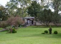 Parterowy budynek z ciemnym dachem i wejściem z rampą, otoczony zielonym trawnikiem i drzewami. Widok na ośrodek wypoczynkowy w słoneczny dzień.