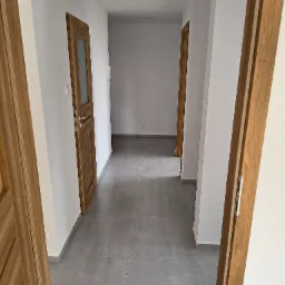 Wykończony korytarz z szarymi płytkami podłogowymi i drewnianymi drzwiami. Świeżo pomalowane białe ściany i widoczne gniazdka elektryczne.