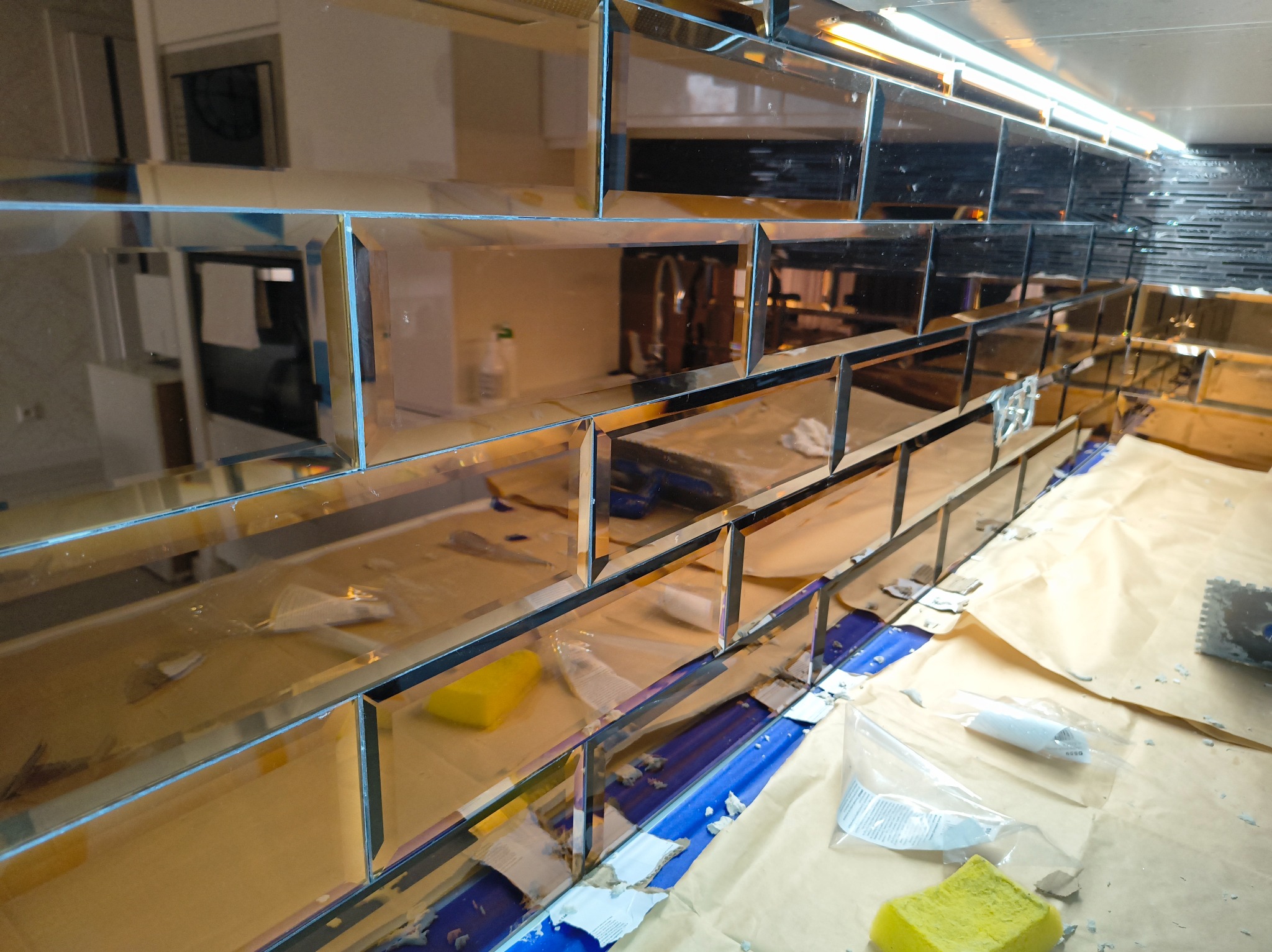 Montaż lustrzanych płytek na ścianie z widocznymi narzędziami i akcesoriami glazurniczymi. Płytki ułożone w rzędach, odbijają otoczenie. Częściowo zakończony proces.