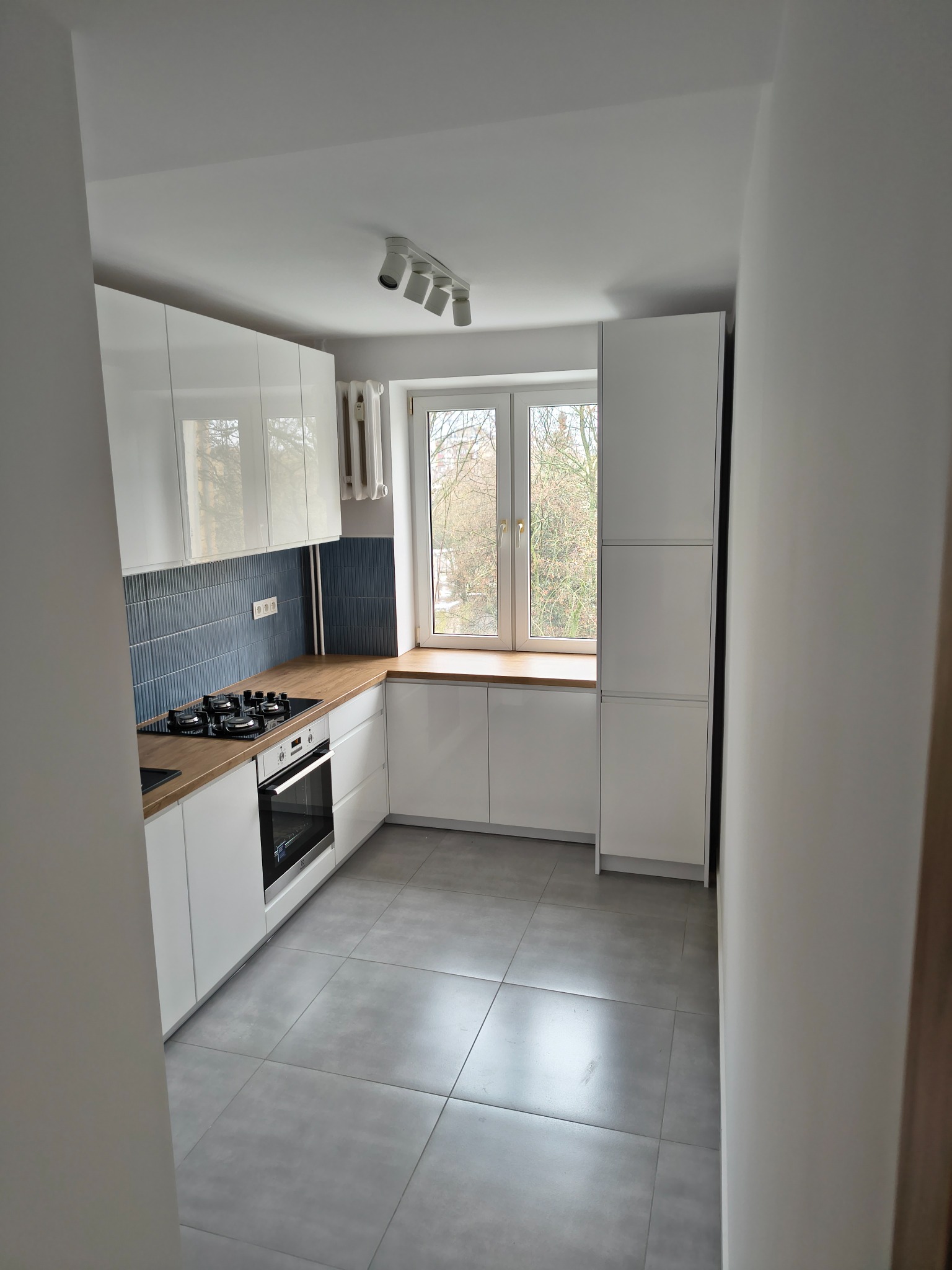 Nowoczesna kuchnia z białymi szafkami, drewnianym blatem i niebieskimi płytkami nad kuchenką. Szare płytki podłogowe i okno z widokiem na drzewa.