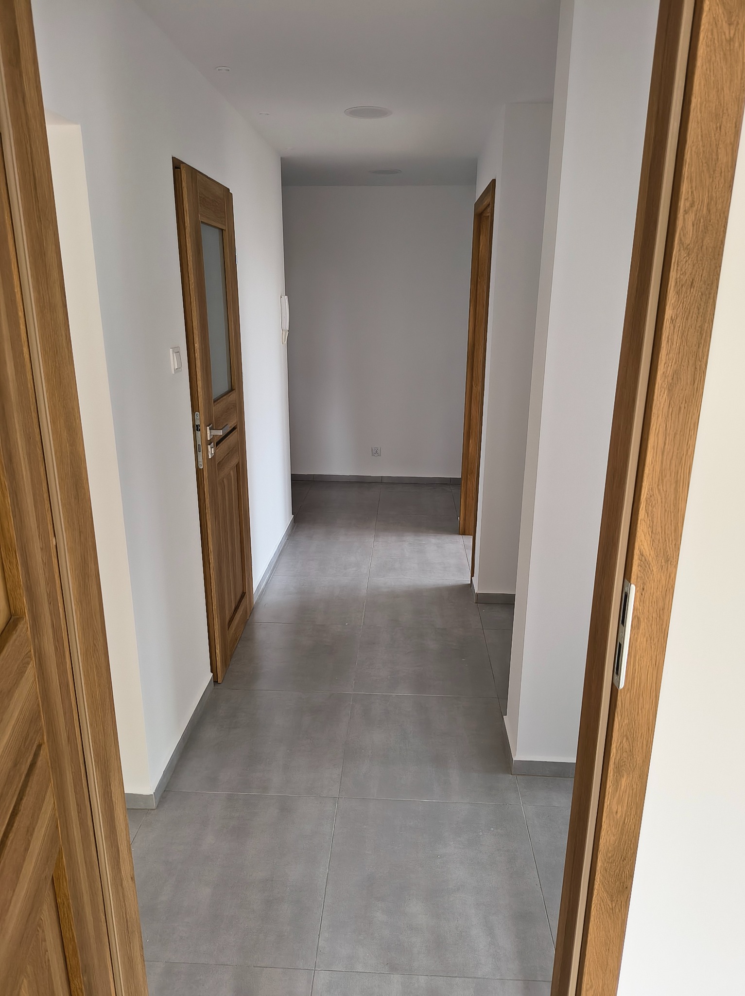 Wykończony korytarz z szarymi płytkami podłogowymi i drewnianymi drzwiami. Świeżo pomalowane białe ściany i widoczne gniazdka elektryczne.