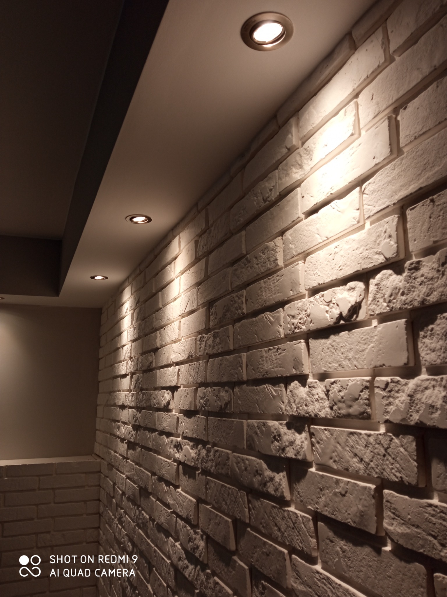 Ściana wykończona białą, dekoracyjną cegłą, oświetlona trzema punktowymi lampami sufitowymi, widok pod kątem.