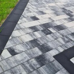 Ułożona szara kostka brukowa z czarnym obramowaniem, widoczna trawa przy krawędzi.