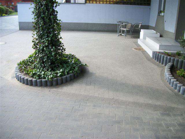 Szara kostka brukowa ułożona w geometryczny wzór, obrzeża z betonowych palisad. Na środku drzewo otoczone kostką, w tle budynek z białymi schodami.