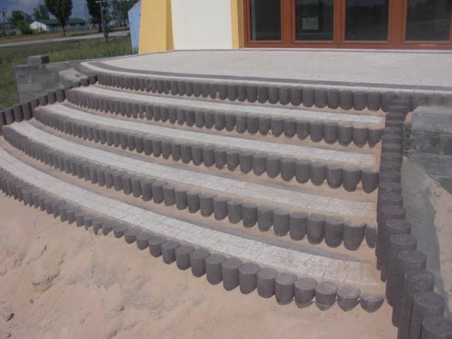 Półokrągłe schody z kostki brukowej w odcieniach szarości i beżu, wykończone obrzeżami z palisady. Widoczny piasek i fragment budynku z oknem.