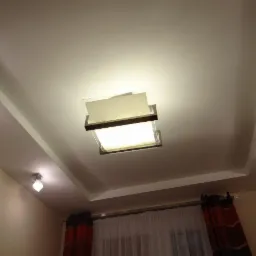 Kaskadowy sufit podwieszany z nowoczesnym oświetleniem centralnym i punktowym. Kremowe ściany i zasłony w tle. Widok z dołu.