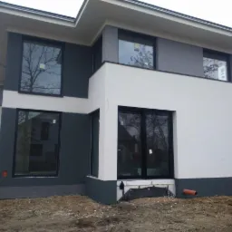 Nowoczesny dom z elewacją w odcieniach szarości i bieli, czarne ramy okienne, widoczne prace wykończeniowe przy gruncie, rura odpływowa.