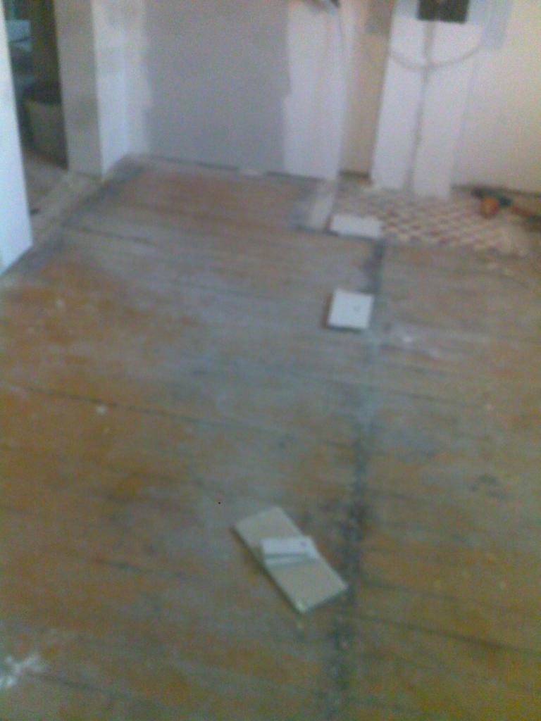 Przygotowanie podłoża pod układanie parkietu: widoczne fragmenty starej podłogi, częściowo odkryte, z pozostawionymi płytkami i materiałami budowlanymi, ściany w trakcie prac wykończeniowych.