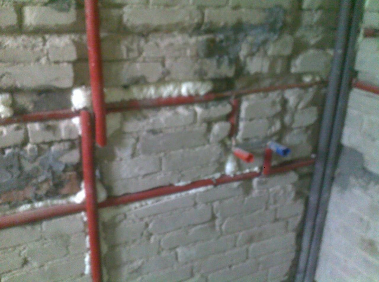 Czerwone rury instalacji wodnej przymocowane do ceglanej ściany z użyciem pianki montażowej, widoczne fragmenty instalacji z rurą czerwoną i niebieską.