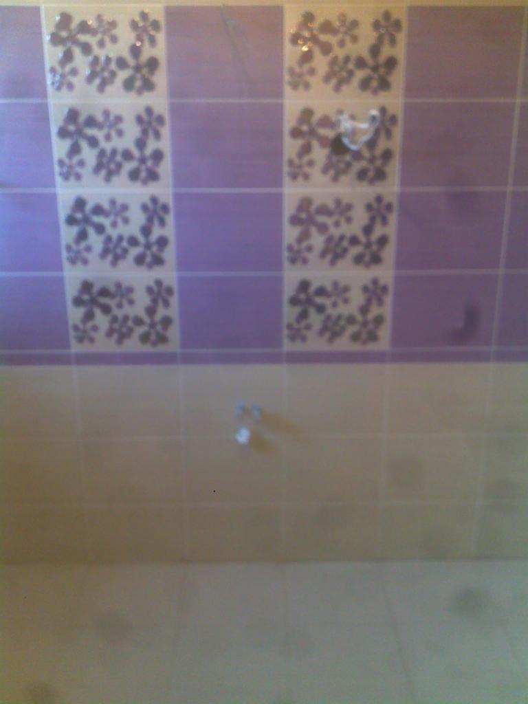 Ściana łazienki wyłożona jasnymi i fioletowymi płytkami z motywem kwiatowym, widoczne elementy armatury.