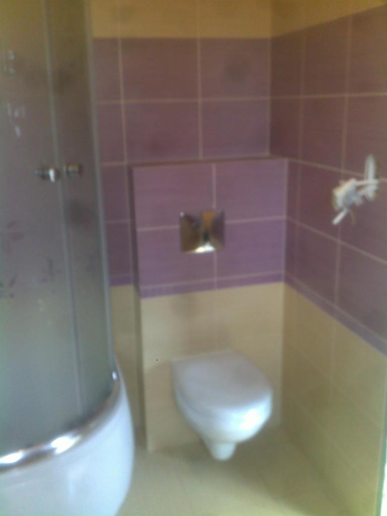 Wykończona łazienka z kabiną prysznicową i podwieszaną toaletą, ściany wyłożone płytkami w odcieniach fioletu i beżu.