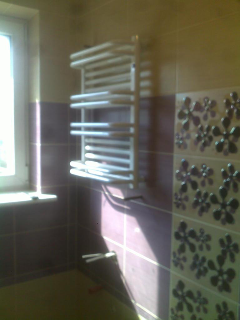 Łazienka z fioletowymi płytkami, grzejnikiem drabinkowym i wzorem kwiatowym na ścianie, oświetlona promieniami słońca wpadającymi przez okno.