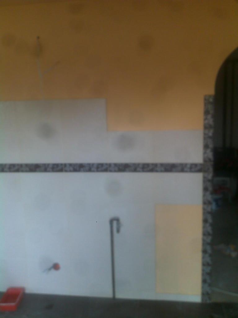 Ściana w trakcie remontu: częściowo położone białe płyty, widoczny fragment dekoracyjnego pasa, rura wystająca ze ściany, czerwone wiaderko na podłodze, żółta ściana w górnej części.