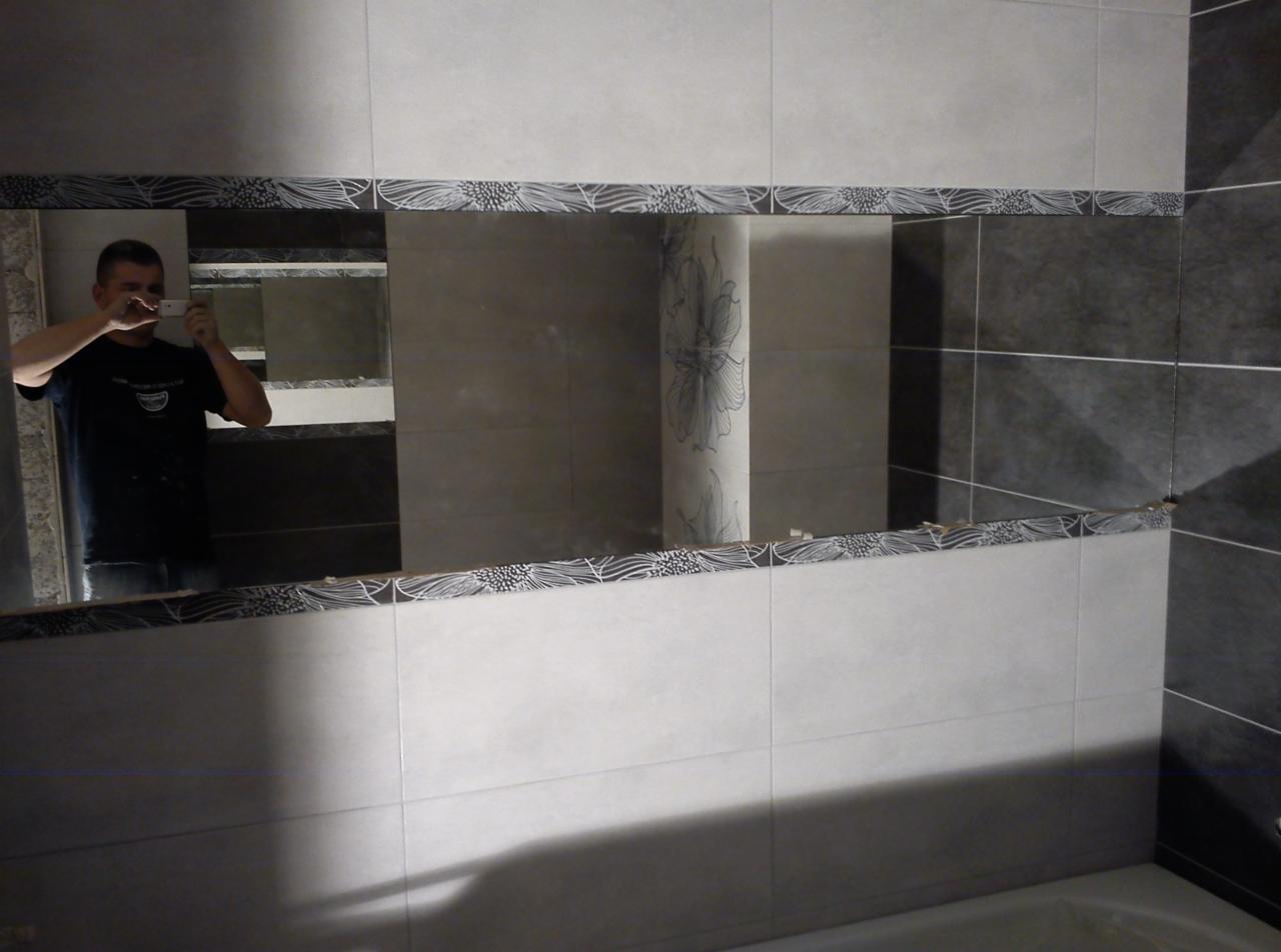 Wykończona ściana łazienki z lustrem w ozdobnej ramie z płytek. W lustrze odbija się osoba robiąca zdjęcie telefonem.