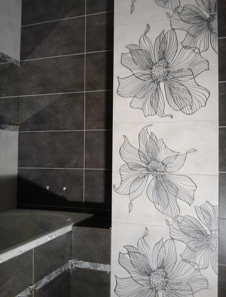 Wykończona łazienka z ciemnymi płytkami i dekoracyjnym panelem z motywem kwiatowym. Widoczna wanna obłożona płytkami z delikatnym wzorem. Minimalistyczny design.