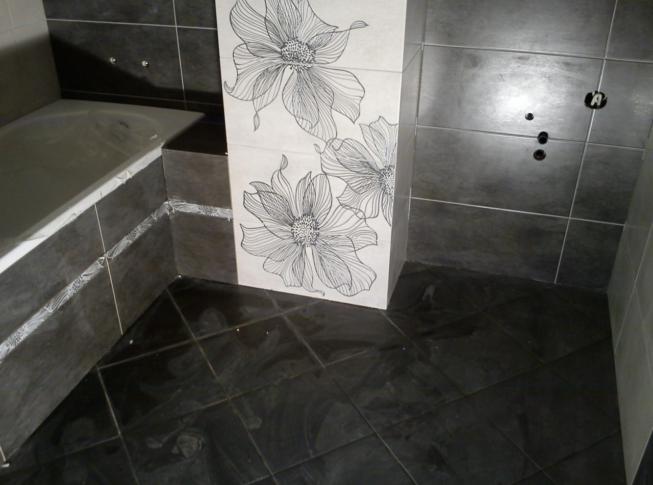 Wykończona łazienka z czarną podłogą, szarymi płytkami ściennymi i dekoracyjnym panelem z motywem kwiatowym. Widoczna wanna i instalacja wodna.