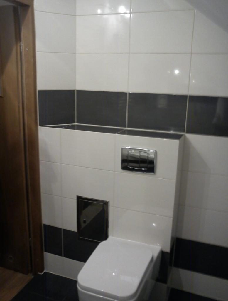 Biała toaleta podwieszana z chromowanym przyciskiem spłukującym na tle ściany wyłożonej białymi i grafitowymi płytkami. Drzwi wejściowe po lewej stronie.