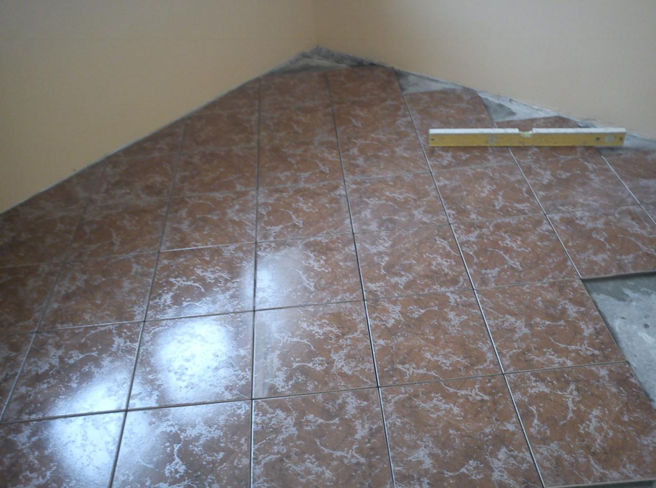 Układanie brązowych, wzorzystych płytek w rogu pomieszczenia, częściowo ukończone. Poziomica leży na płytkach. Widoczny fragment surowej podłogi.