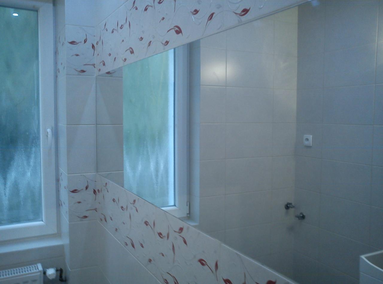 Wykończona łazienka z białymi płytkami i dekoracyjnym pasem z motywem liści. Widoczne okno i odbicie fragmentu łazienki w lustrze.