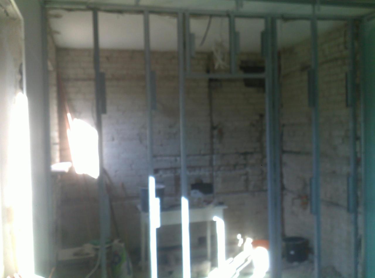 Stelaż z profili metalowych przed murowaną ścianą w trakcie budowy lub remontu, widoczne przewody elektryczne.