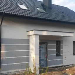 Nowoczesny dom jednorodzinny z szarą elewacją, białymi pasami dekoracyjnymi i ciemną dachówką, widoczne okna dachowe i częściowo zakryte wejście folią ochronną.