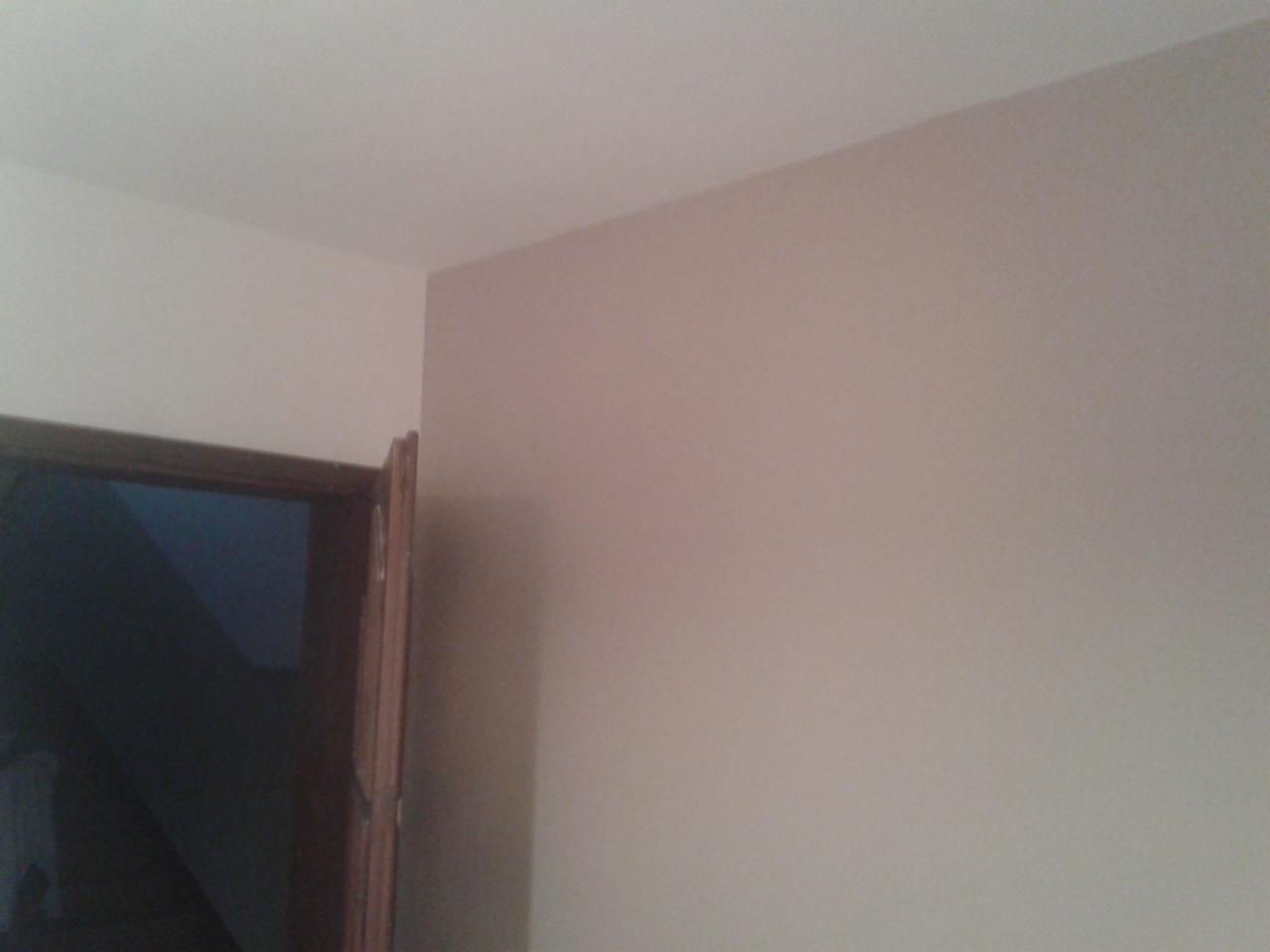 Pomalowany narożnik pokoju w odcieniach beżu, z widocznym fragmentem drewnianej ościeżnicy drzwi. Gładkie ściany i sufit. Neutralne oświetlenie.