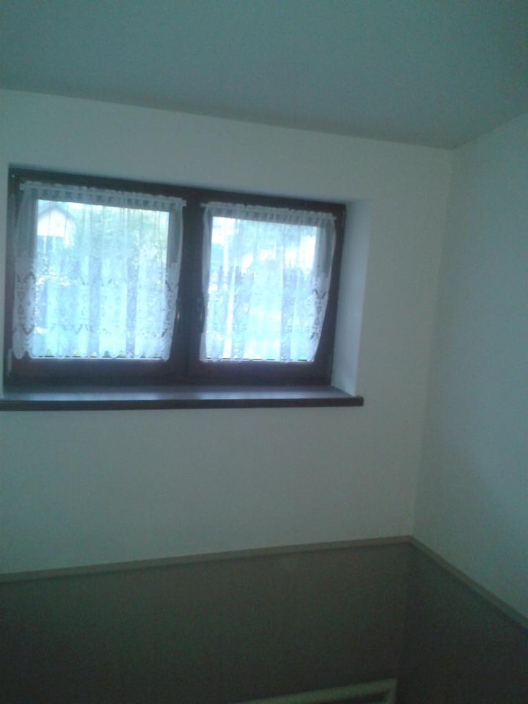 Wnętrze po malowaniu: Ściany i sufit pomalowane na biało, okno z brązową ramą i firankami. Podłoga w odcieniach szarości. Widok na fragment odnowionego pokoju.