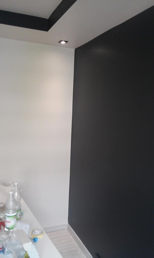 Dwukolorowe wnętrze po malowaniu: ściana w głębokim odcieniu grafitu kontrastuje z bielą, widoczna listwa przypodłogowa, na blacie roboczym butelki z wodą i taśma malarska.