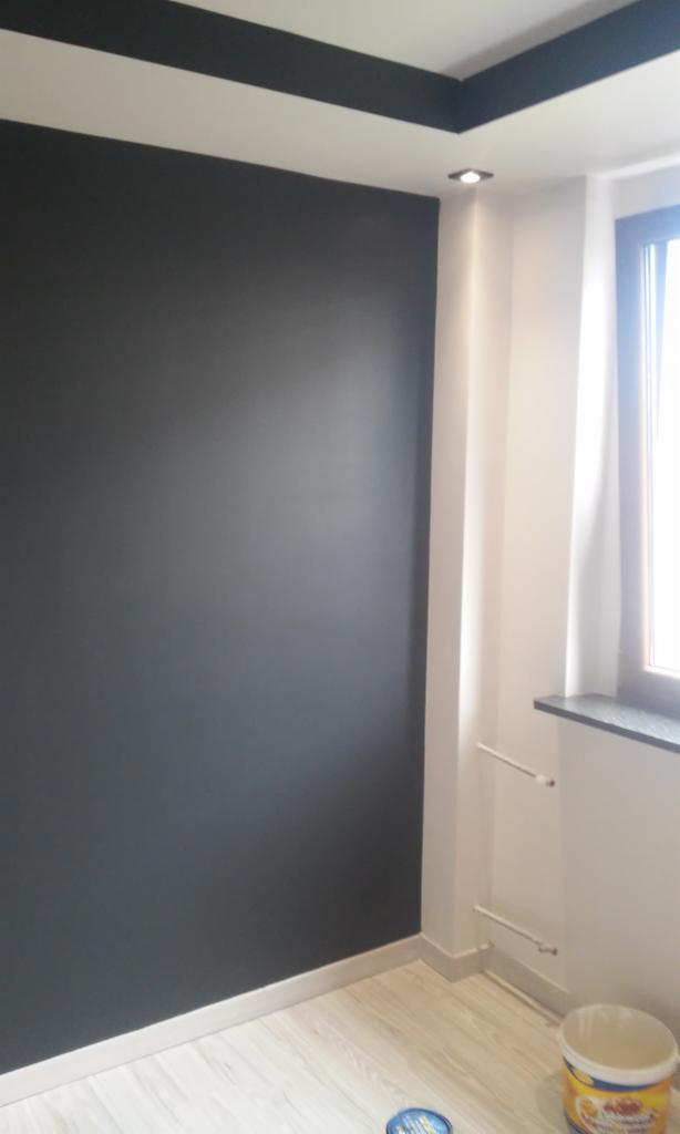 Ściana pomalowana na ciemny grafit z czarnym pasem przy suficie, widoczny fragment okna z zasłoną i puszka po farbie na podłodze.