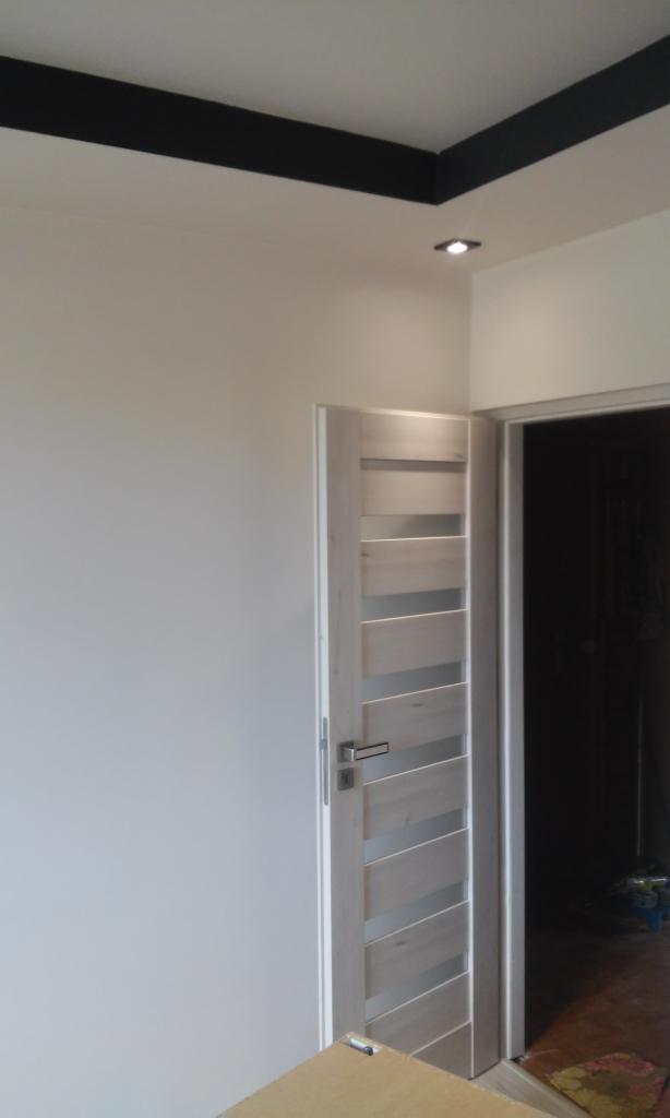 Wnętrze po odświeżeniu: Białe ściany z czarnym pasem pod sufitem, nowoczesne drzwi wewnętrzne z przeszkleniami i widoczne oświetlenie punktowe.