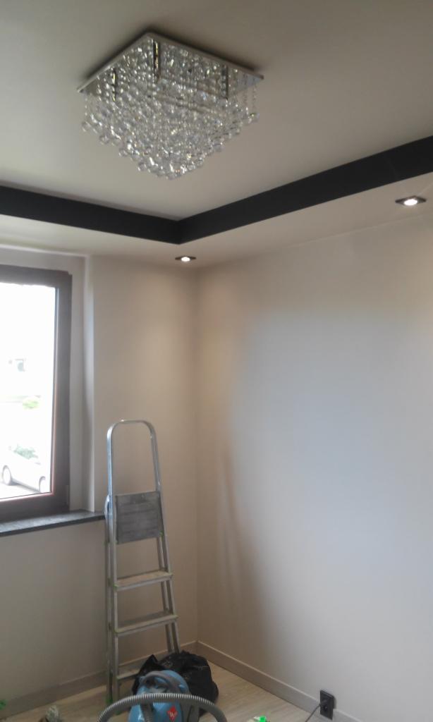 Wykończone wnętrze z kryształowym żyrandolem, czarnymi listwami sufitowymi i aluminiową drabiną opartą o ścianę przy oknie.