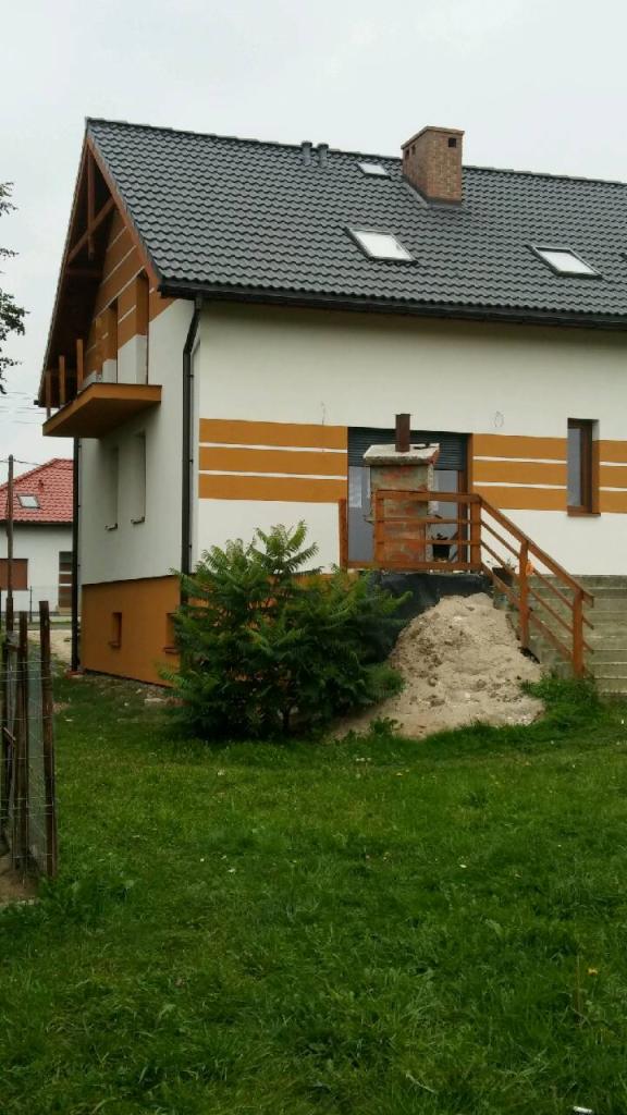 Dwukondygnacyjny dom z ciemnoszarą dachówką, kremową elewacją z poziomymi, brązowymi pasami, balkonem i schodami wejściowymi, częściowo zasłonięty krzewem i stertą piasku.