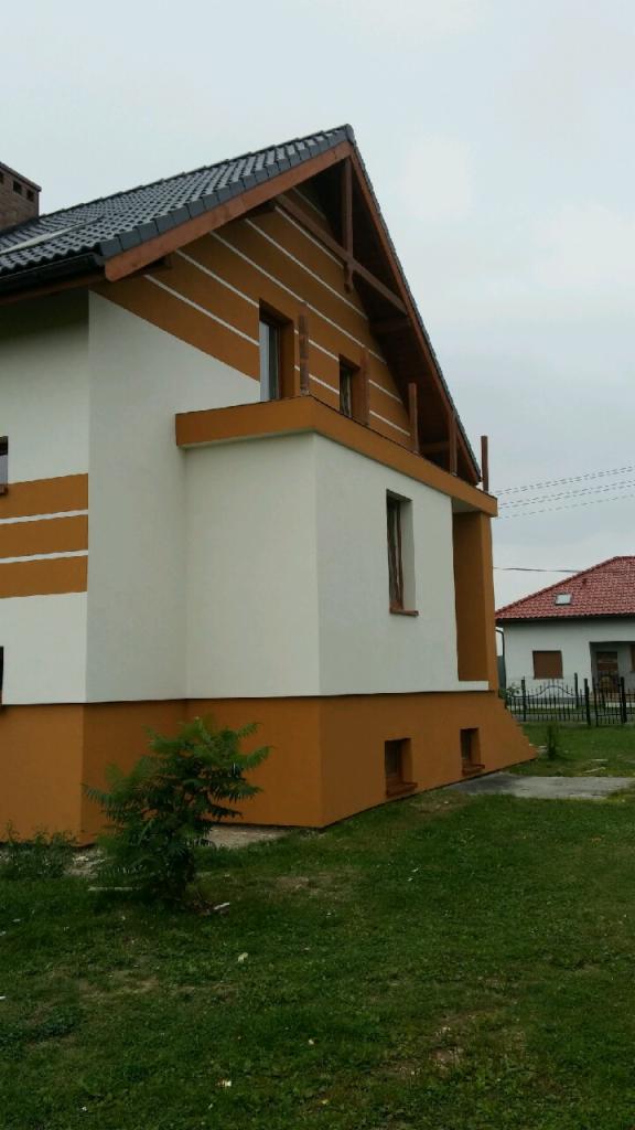 Dwukolorowa elewacja domu jednorodzinnego w Studzionce, z wyraźnym podziałem na część białą i pomarańczową, oraz pasami na górnej części ściany szczytowej, widok z boku na tle pochmurnego nieba.